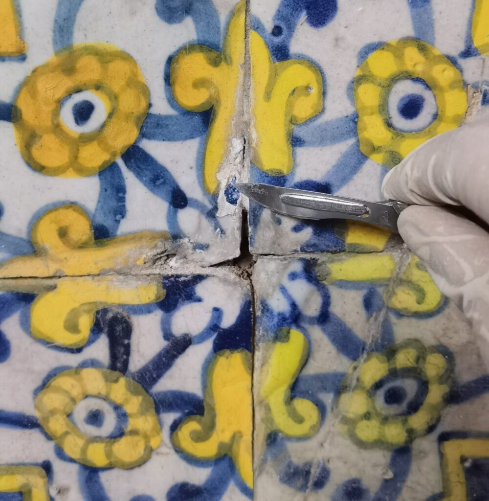 IMG_20230807_141951_resized_20251030_101733079 Limpeza mecânica controlada de juntas entre azulejos históricos, utilizando bisturi de precisão para remoção de sujidades e argamassas deterioradas. Esta fase do restauro permite restabelecer a integridade visual e estrutural das peças, preparando a superfície para a reintegração cromática e conservação final do património azulejar.